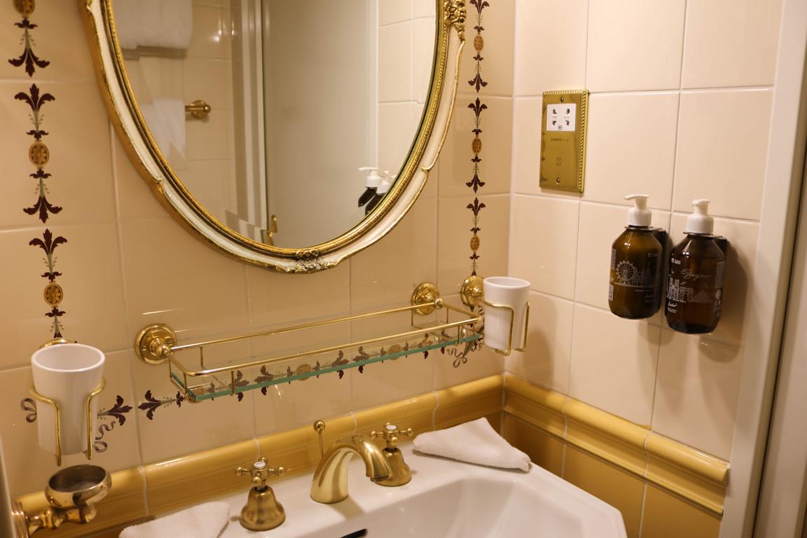 Das im Belle-Époque-Stil gestaltete Duschbad fügt sich mit kunstvoll gestalteten Porzellanwaschtisch, und passenden Zahnputzbechern mit goldfarbenden Wasserhahn und Spiegel harmonisch in das historische Schiffskonzept ein
